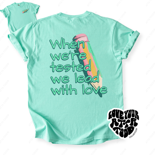 Painted pencil graphic with encouragement text “When We Are Tested We Lead With Love,” thick paint texture and high-contrast color, printed on a Comfort Colors® tee.