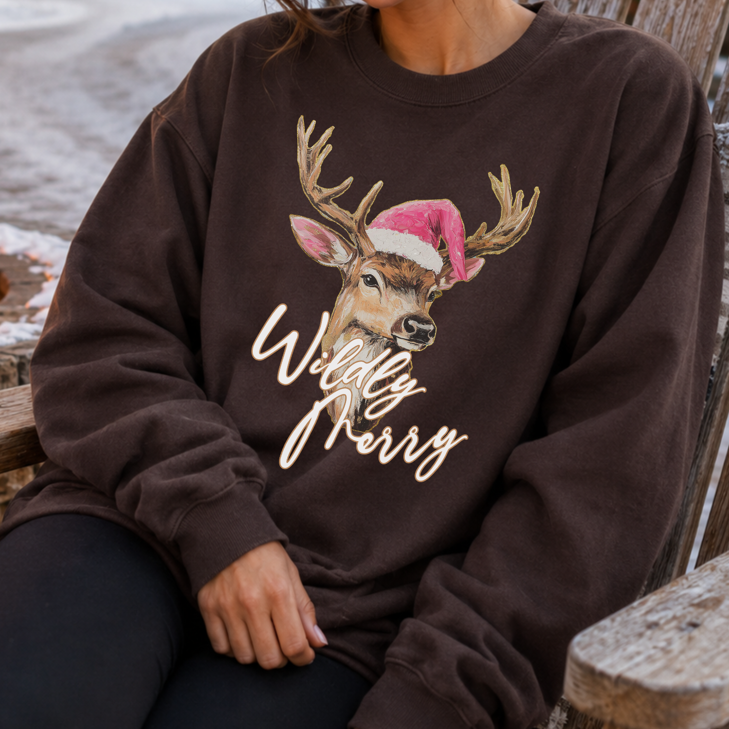 A person wearing a dark brown sweatshirt with a graphic of a deer wearing a pink Santa hat and the words 'Wildly Merry' in white script, seated on a wooden bench outdoors.