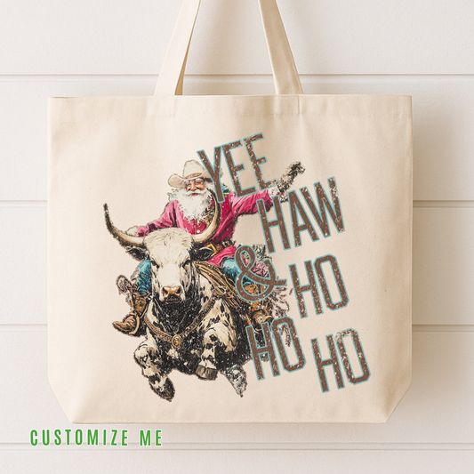 A cream-colored canvas tote bag with a rustic graphic of a cowboy riding a bucking bull, overlaid with the text 'YEE HAW & HO HO HO'. The bag is displayed against a white wood-paneled wall, with a green 'CUSTOMIZE ME' text overlay at the bottom left.