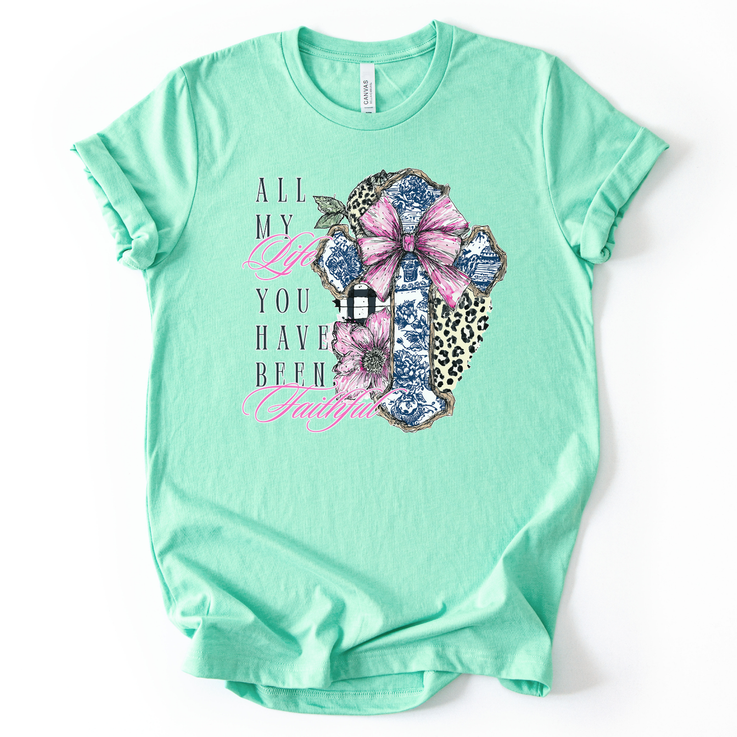 This trending graphic print is a decorative cross collage made up of blue toile-style pattern pieces, leopard print accents, and a black-and-white buffalo check strip, topped with a large pink bow.  On a heather mint t-shirt