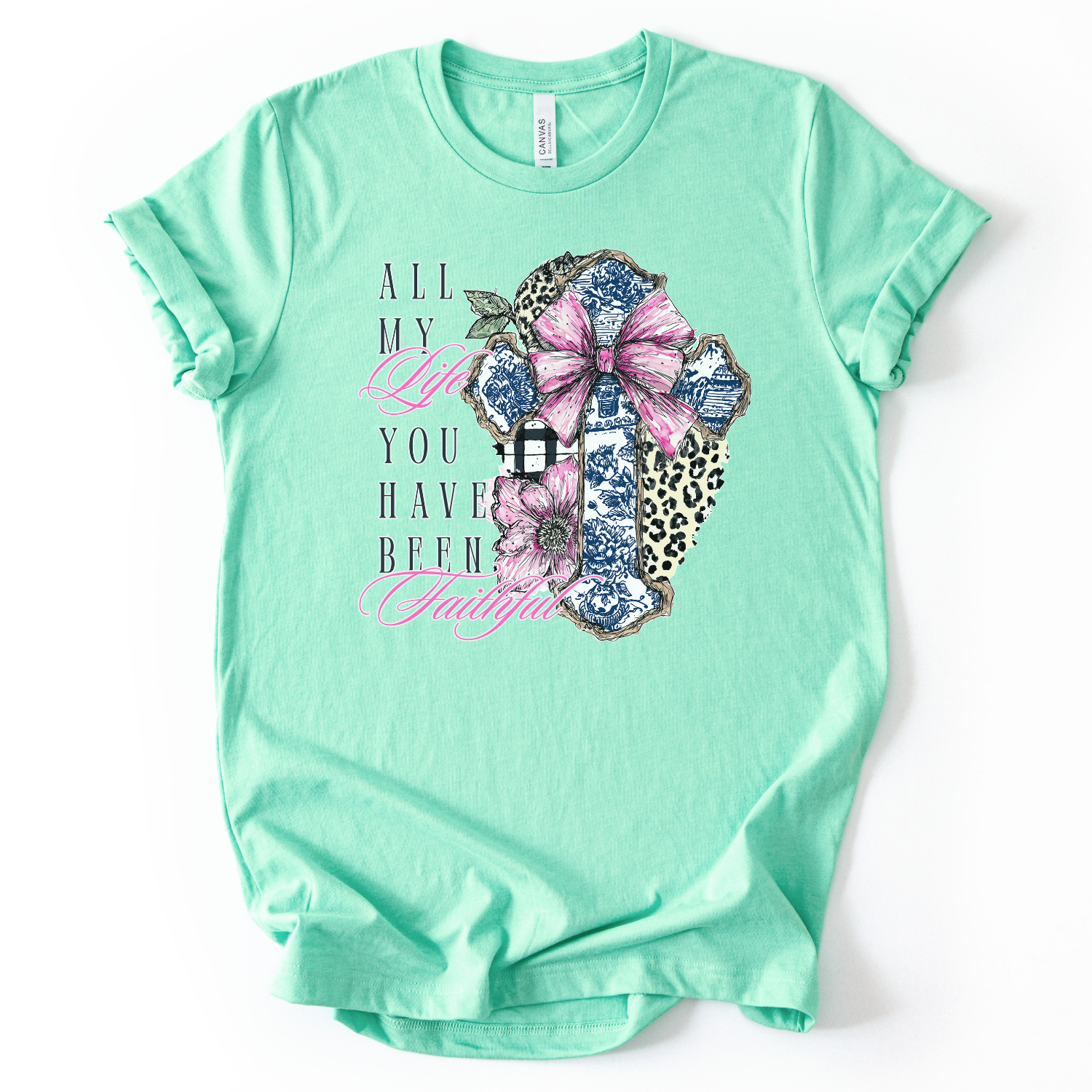 This trending graphic print is a decorative cross collage made up of blue toile-style pattern pieces, leopard print accents, and a black-and-white buffalo check strip, topped with a large pink bow.  On a heather mint t-shirt