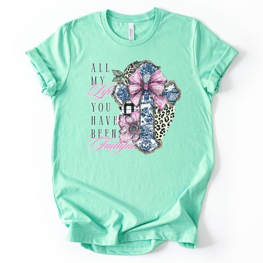 This trending graphic print is a decorative cross collage made up of blue toile-style pattern pieces, leopard print accents, and a black-and-white buffalo check strip, topped with a large pink bow.  On a heather mint t-shirt